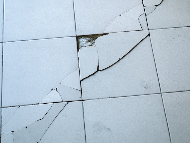 Close up photo of light gray tile with large diagonal cracks and chips.